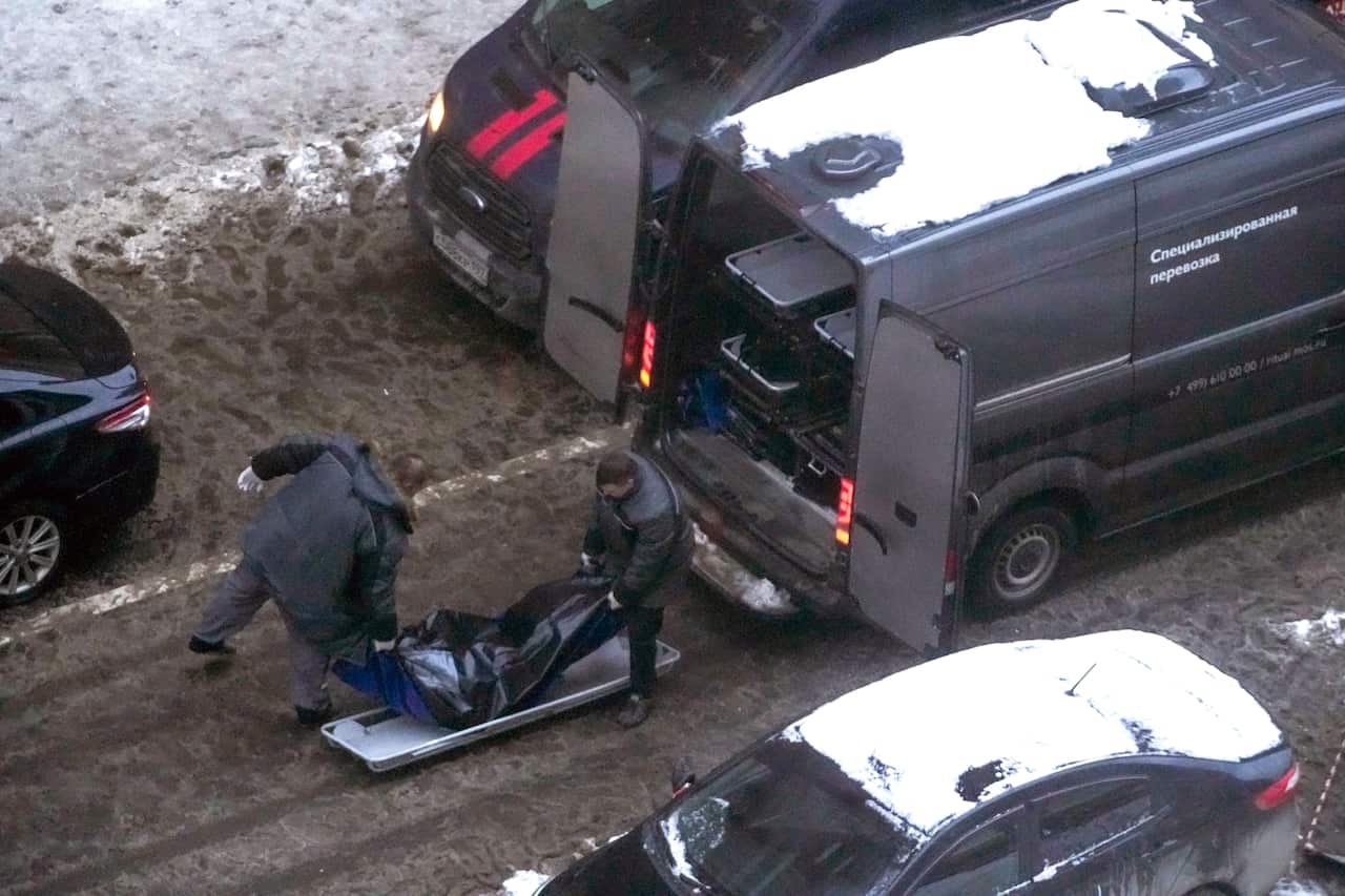 A body bag is being lifted into a van by two men in black clothes.