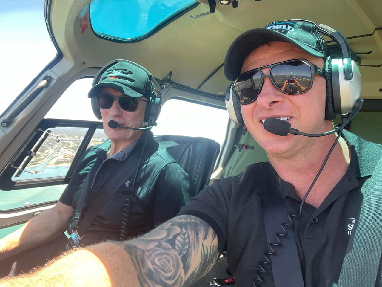 Two men in black shirts, sunglasses and headsets in a helicopter. 