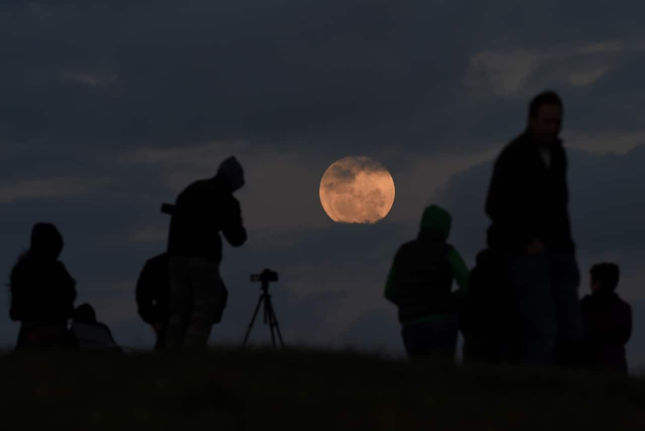 An orange moon in a night sky with silhouettes of people and a telescope in the foreground. 