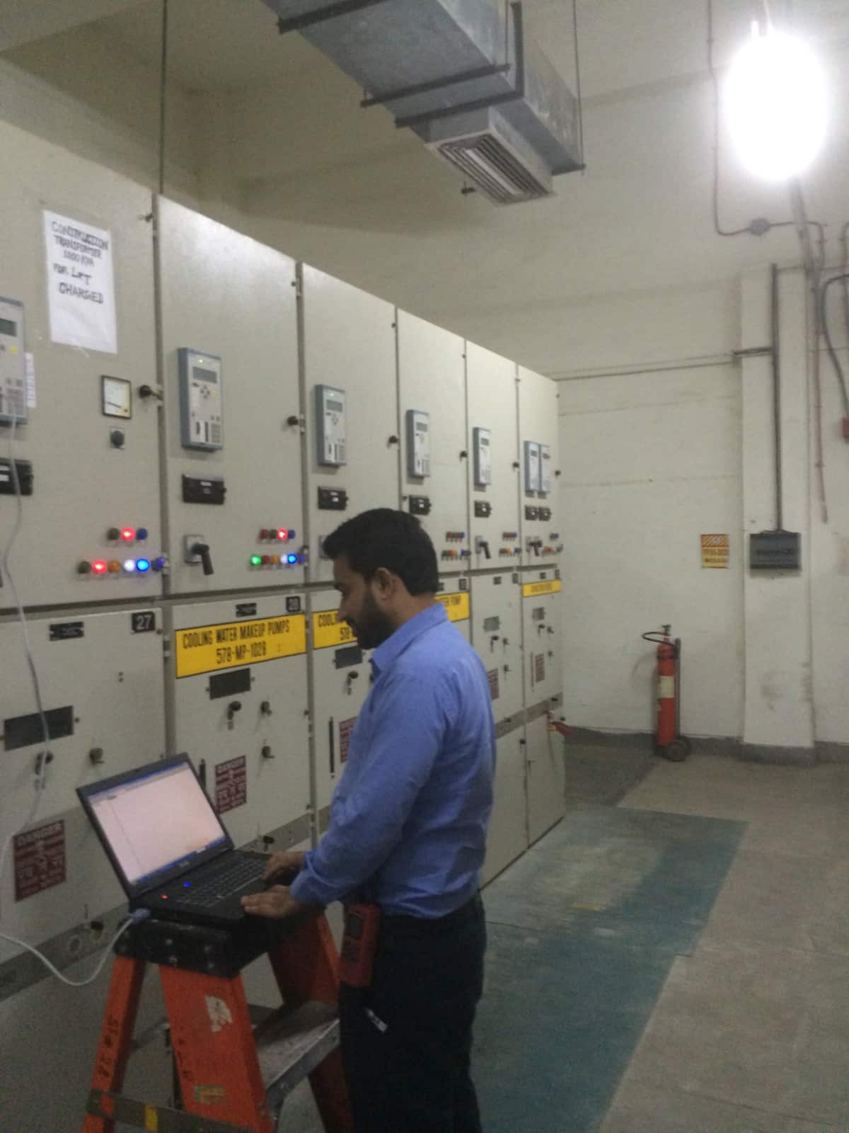A man in a blue shirt works on a laptop inside an industrial mill, with electrical switchboards in front of him.