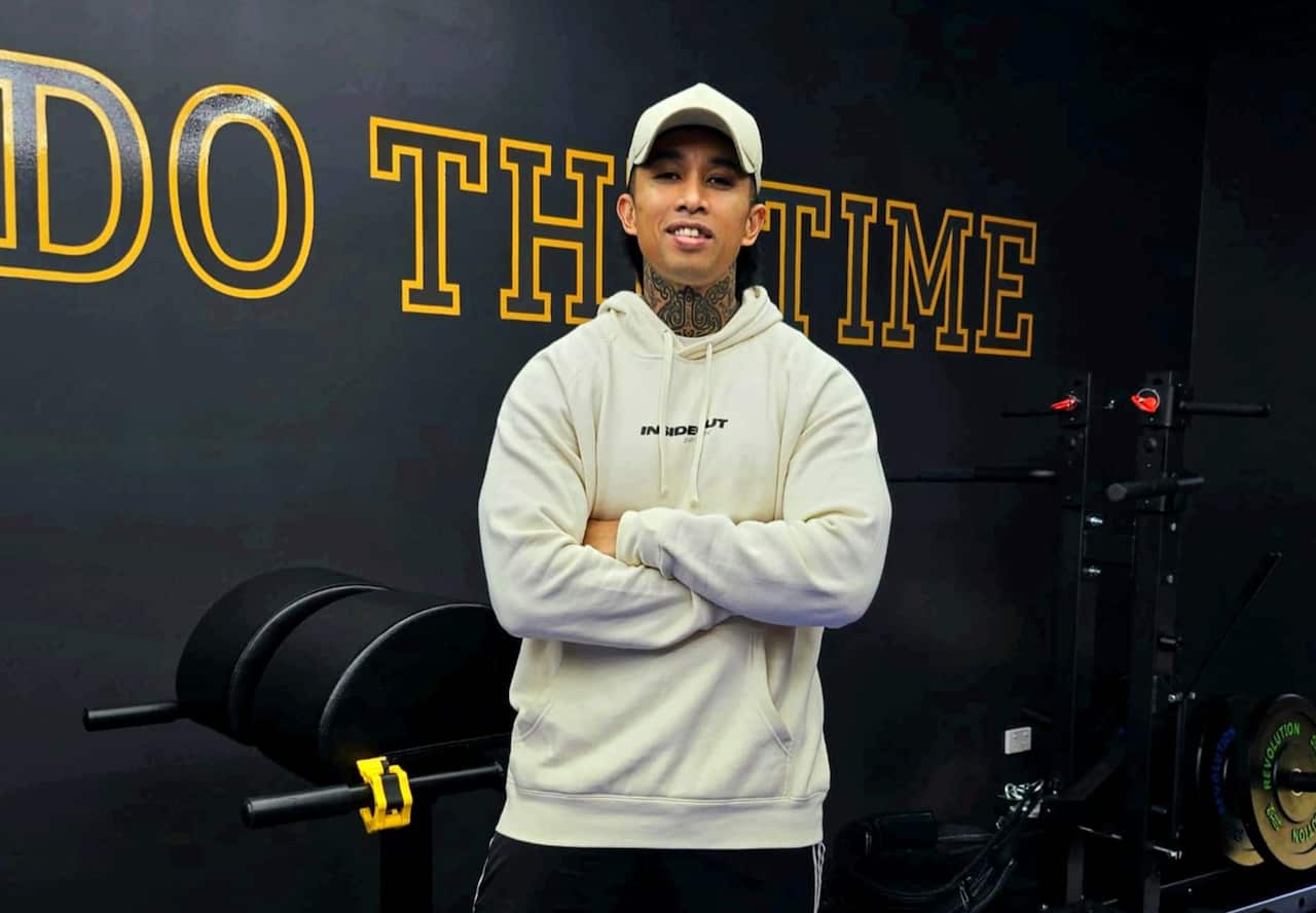 A young man wearing a white hoodie and a cap smiles in a gym.