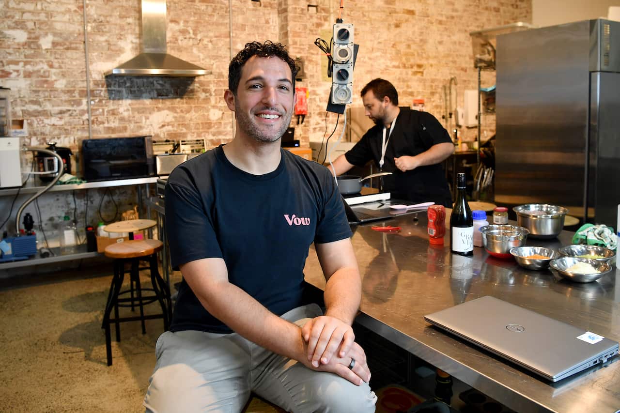 A man is seated on a bar stool while another works in the background.