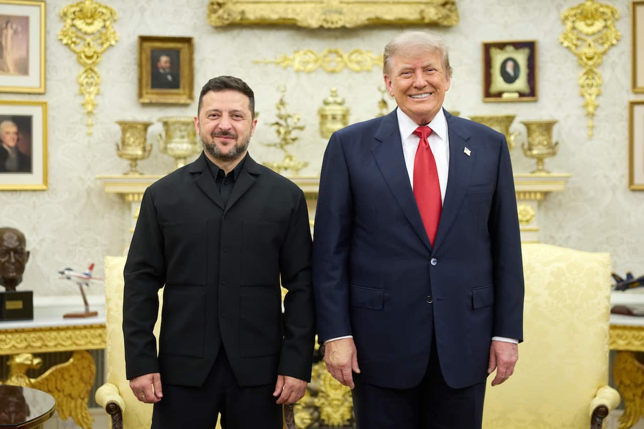 A middle-aged white man in a black suit, standing next to an older white man in a navy suit. Both are smiling. In the background, gold ornaments sit on a wall.