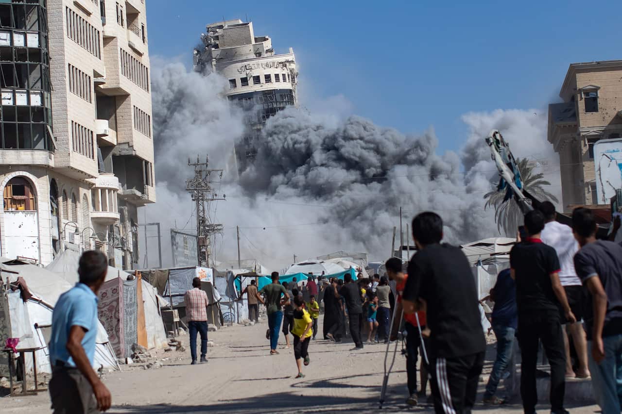 Smoke surrounds a building that is collapsing, as onlookers watch from the street below.