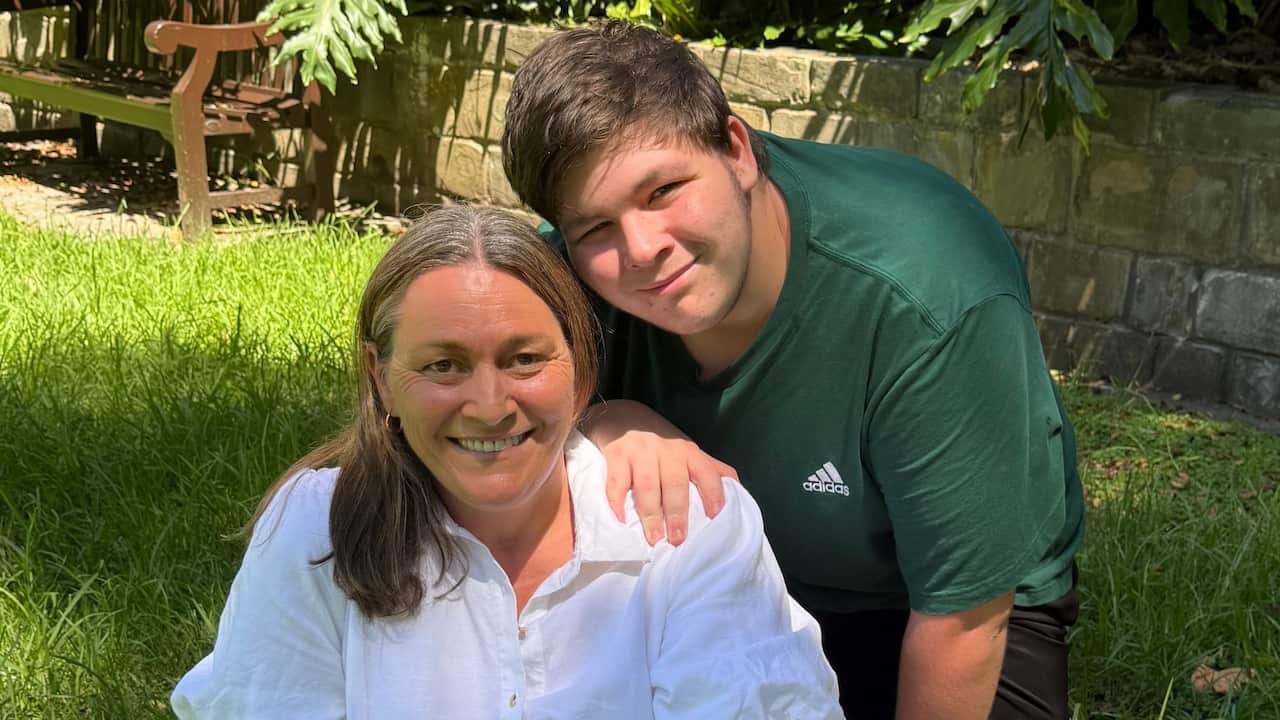 a middle aged woman and her teenage son smile together sitting on the grass in a park