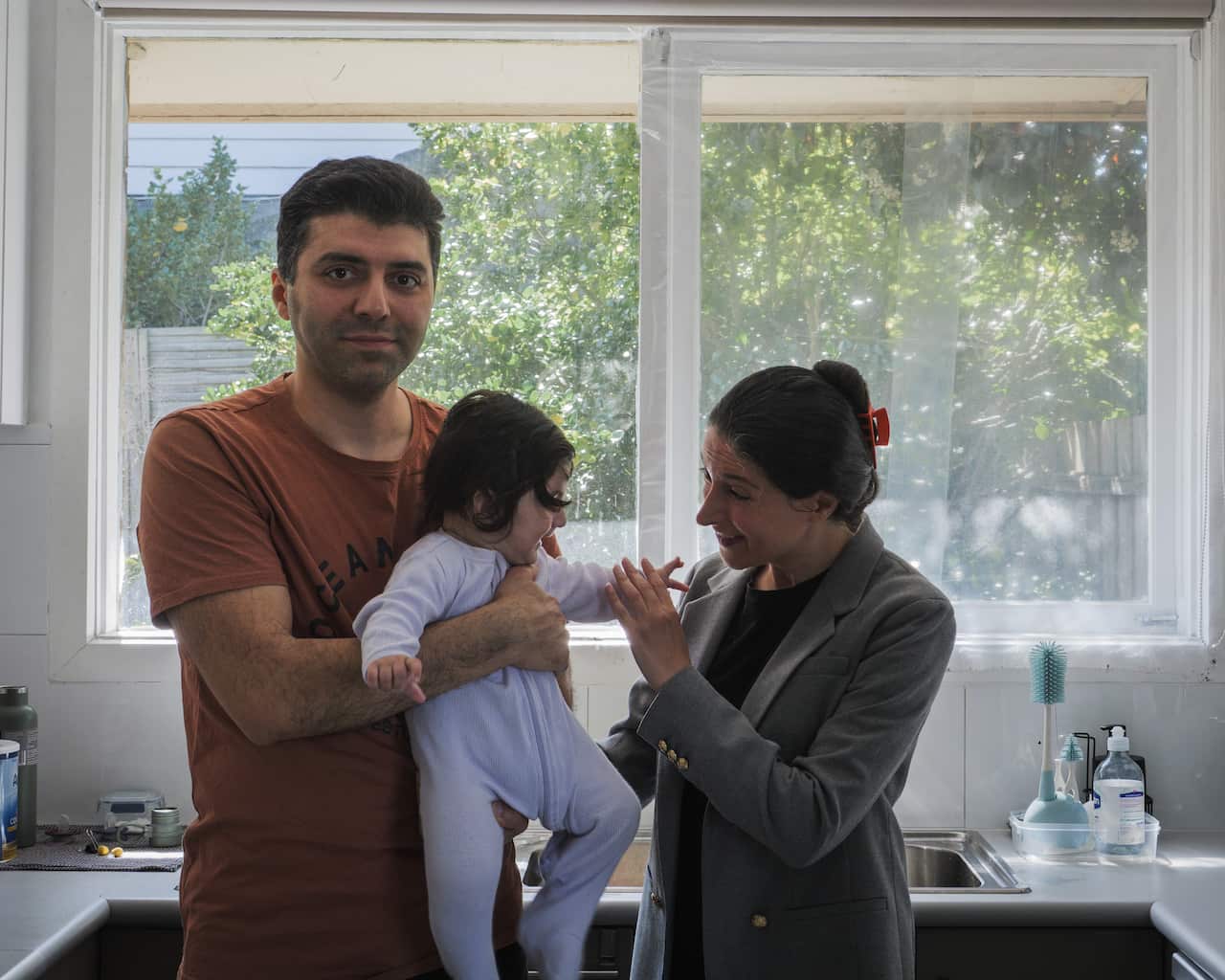 A father holds his young child while standing next to the mother in a kitchen. The family is posing for a photo in front of a window with a view of a backyard.