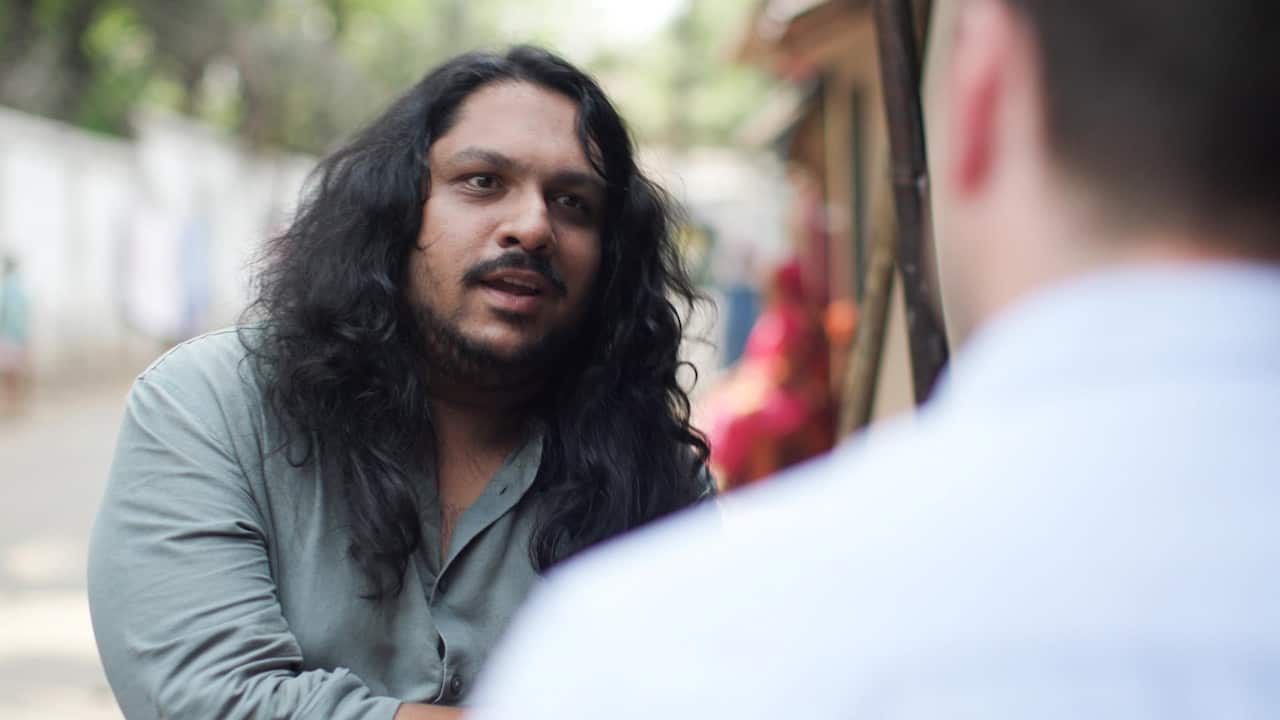 A South Asian man with long curly hair and a beard is talking to another person, who's seen from behind
