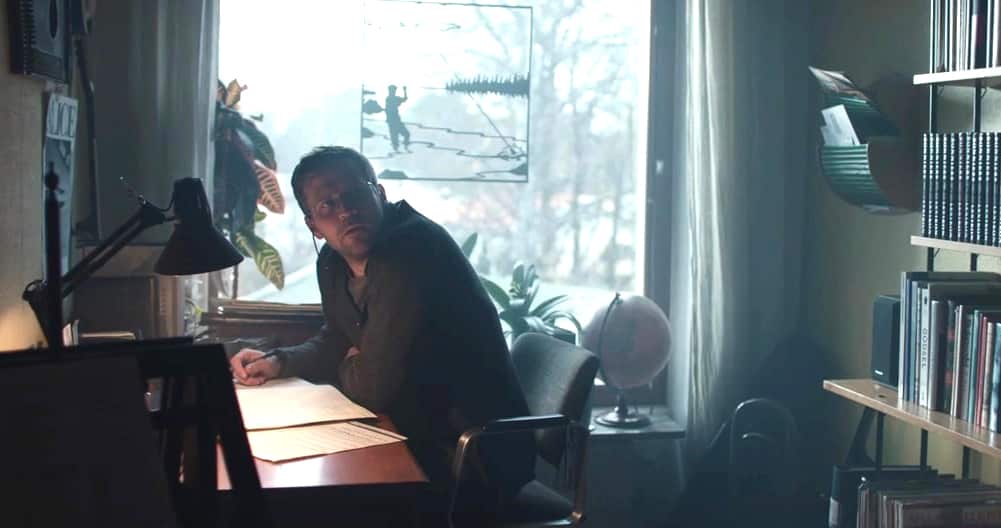 A man in glasses sits working at a desk in a study, beside a window. Trees can be seen outside. His head is turned as if looking at someone.  