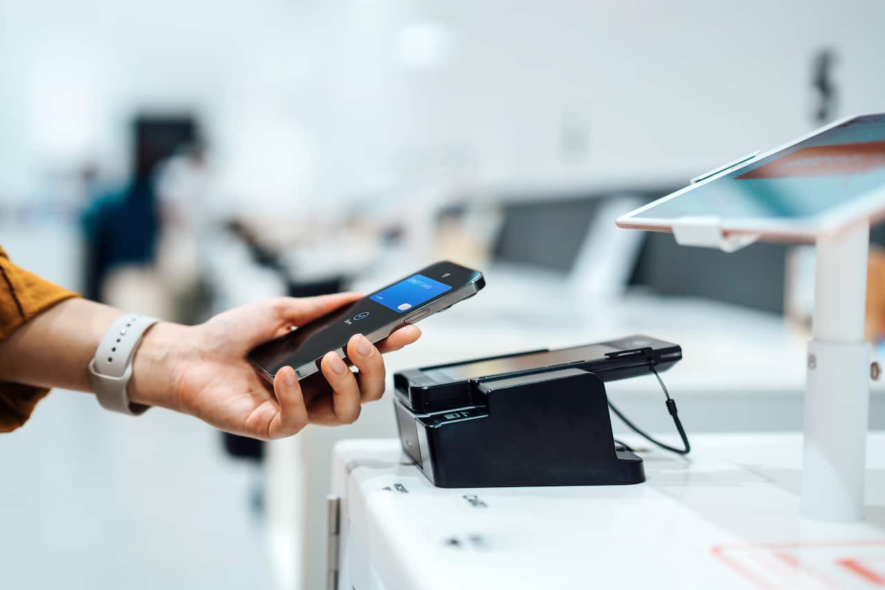 Close-up shot of a female hand holding a smartphone near a credit card machine, making a contactless payment to pay for her shopping at a self-checkout machine in a store.