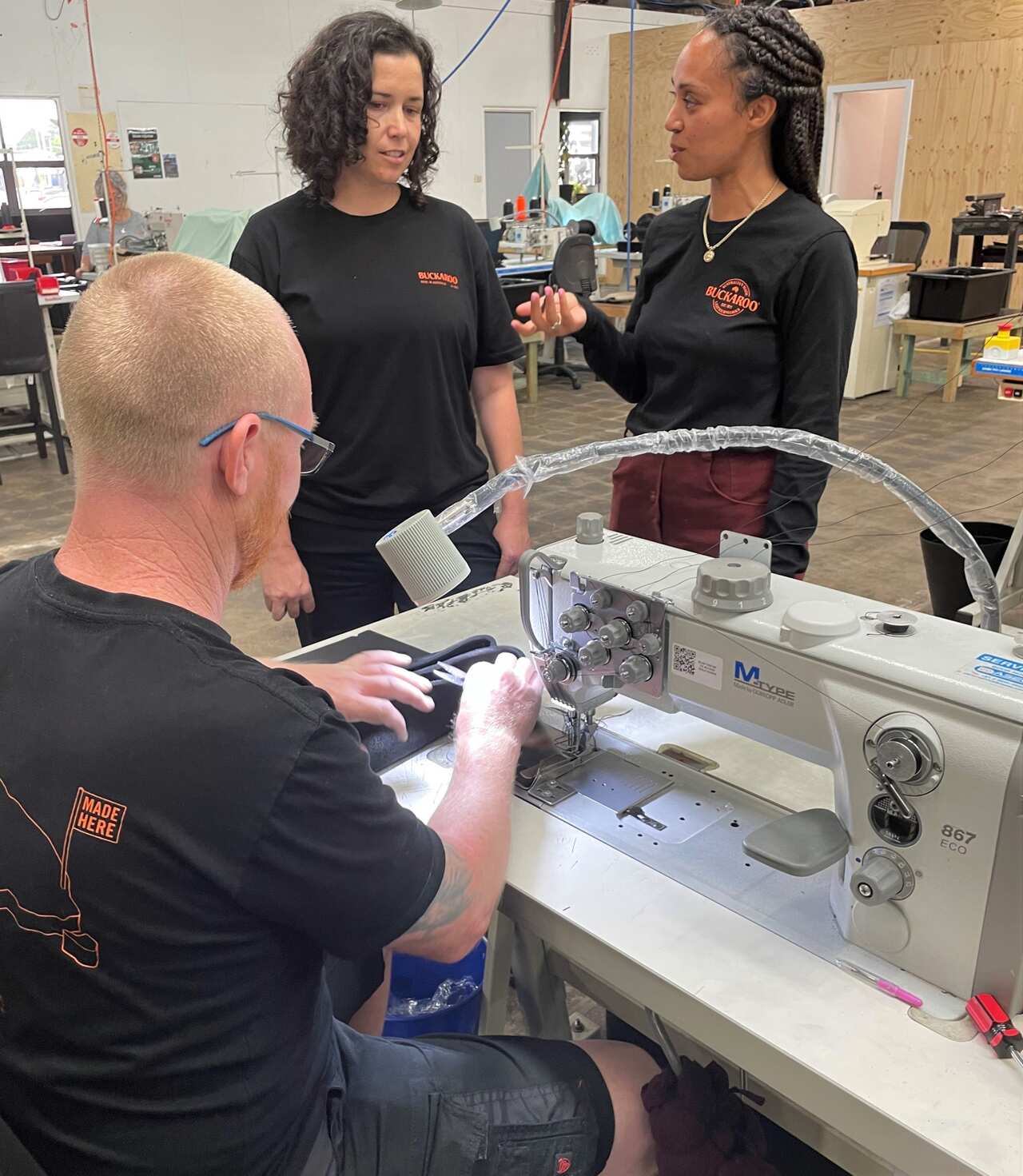 A woman in a black t-shirt stands in a factory talking to workers.