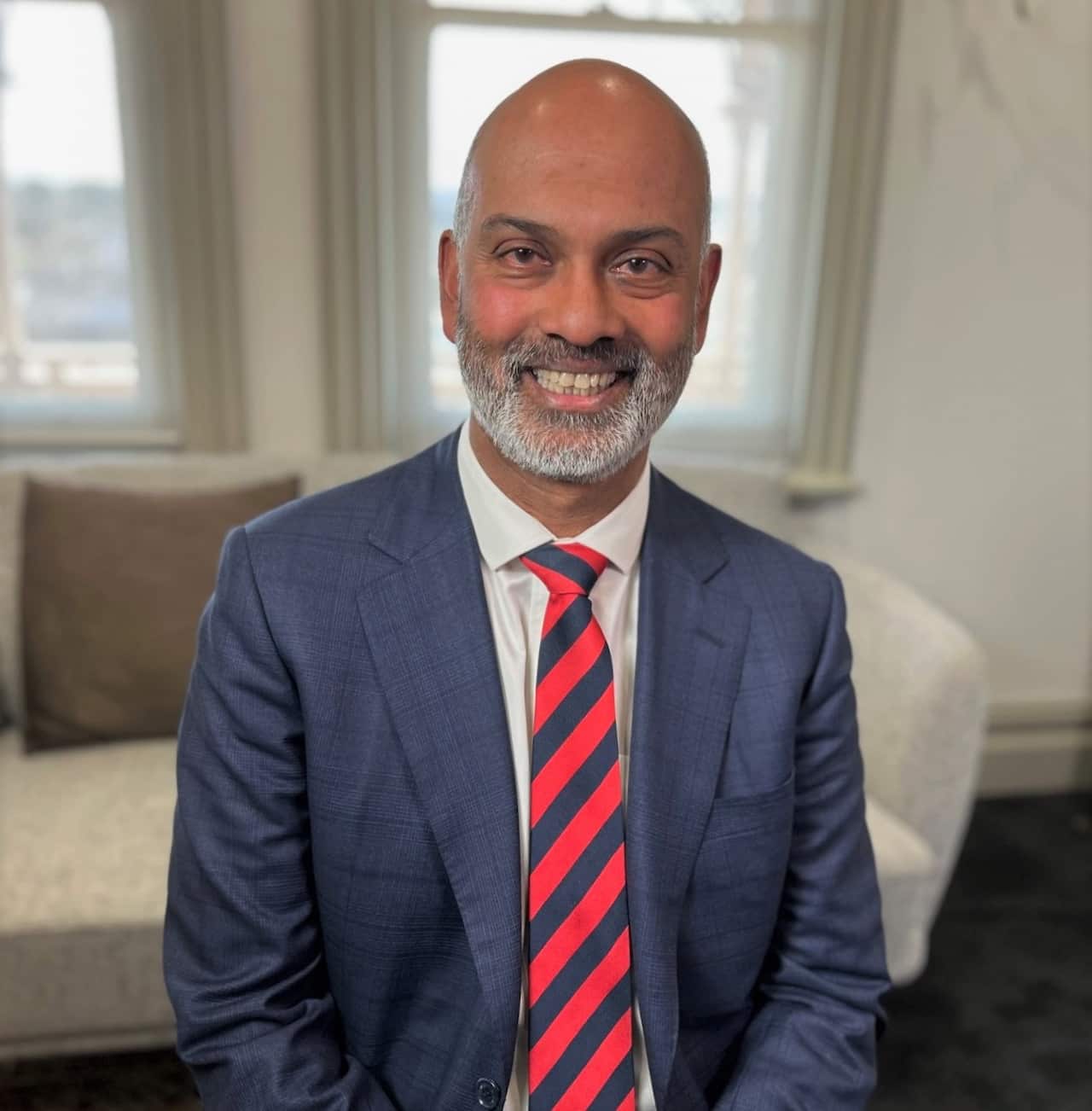 A man in a blue suit with a red striped tie smiles at the camera.