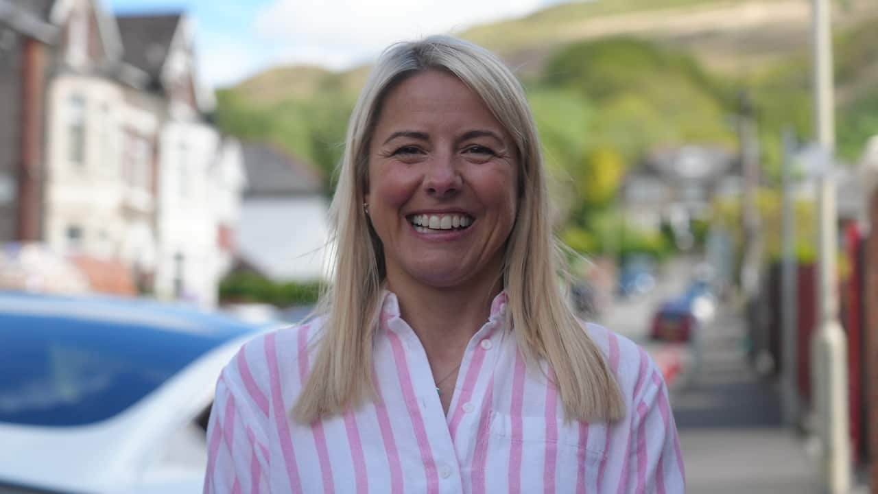 A smiling woman with blonde hair, wearing a pink and white striped shirt, stands on a residential street with houses and a hillside in the background.