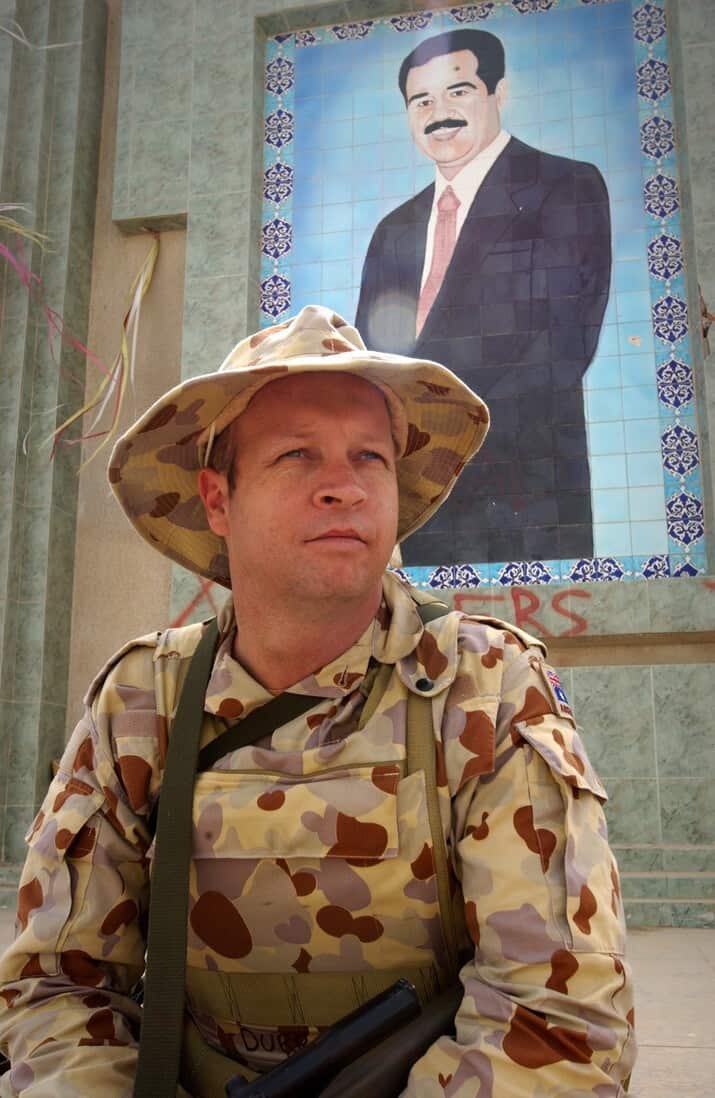 A younger white man in military uniform and hat gazing towards his left. Behind him is a tiled painting of a Middle Eastern man in a suit.