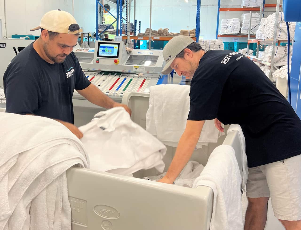 Two men in black t-shirts leaning into a trolley filled with white washing.