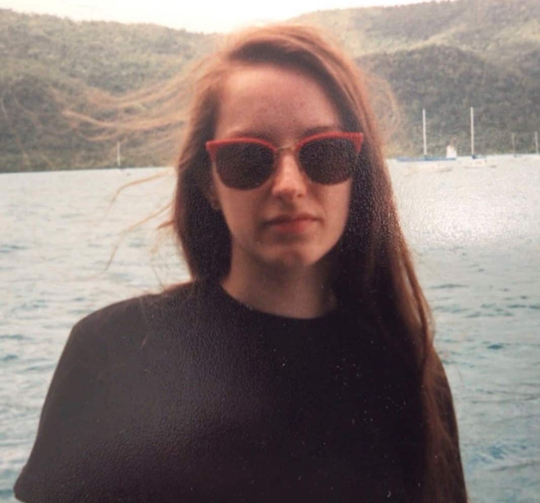 A woman stands outside with the water in the background. She's wearing sunglasses and has a serious expression.
