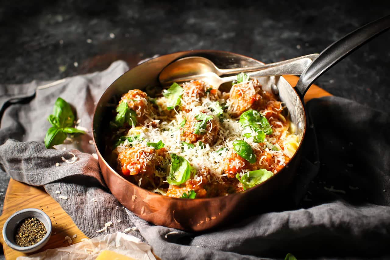 A deep copper saucepan sits on a grey napkin. It holds meatballs and pasta, topped with basil leaves and grated parmesan. 