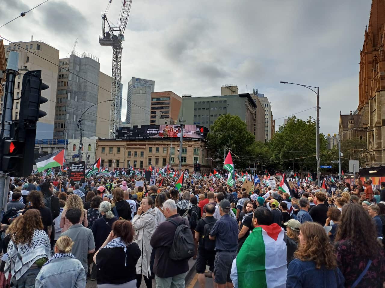 A large crowd of people gathered in an urban intersection carries Palestinian flags and protest signs, set against a backdrop of city buildings and a construction crane under an overcast sky.