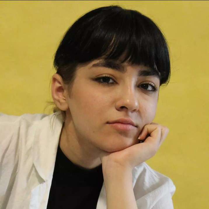 Photo of a young girl with black bangs, posing with her hand on her chin. 