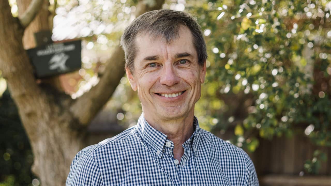a headshot of a middle aged man in a checkered button-up shirt in a backyard
