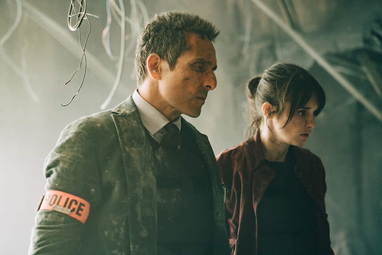 A man in a suit, covered in dust and with an armband that says POLICE, and a woman in a dark jacket stand looking down and to one side. Loose wires hang from the ceiling behind them. 