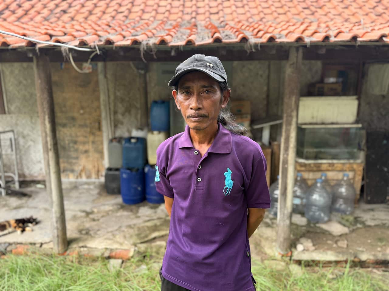 A man wearing a purple polo shirt and grey cap standing outside a single-storey home with a large tiled awning at the front of it