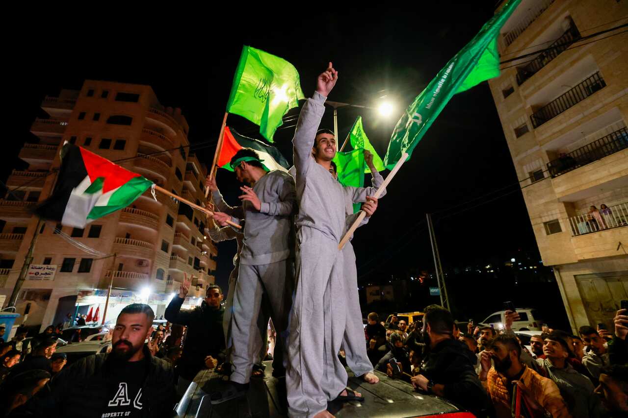 Two men in grey jumpers stand on a car waving flags.