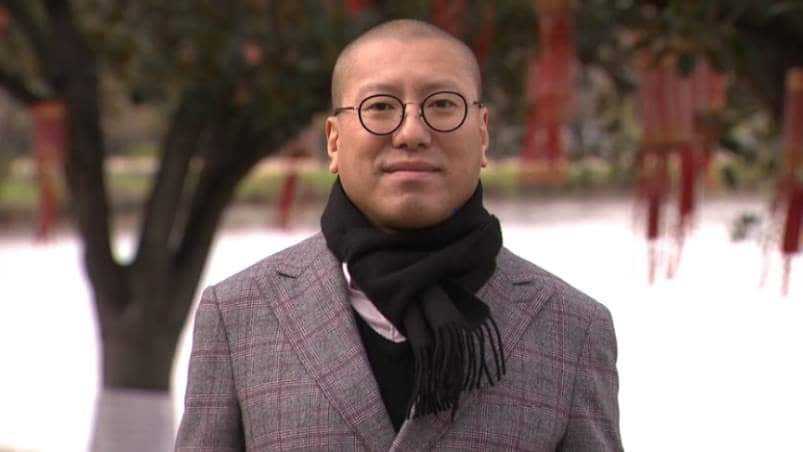 Kevin smiles at the camera, wearing glasses, a checkered blazer and a scarf