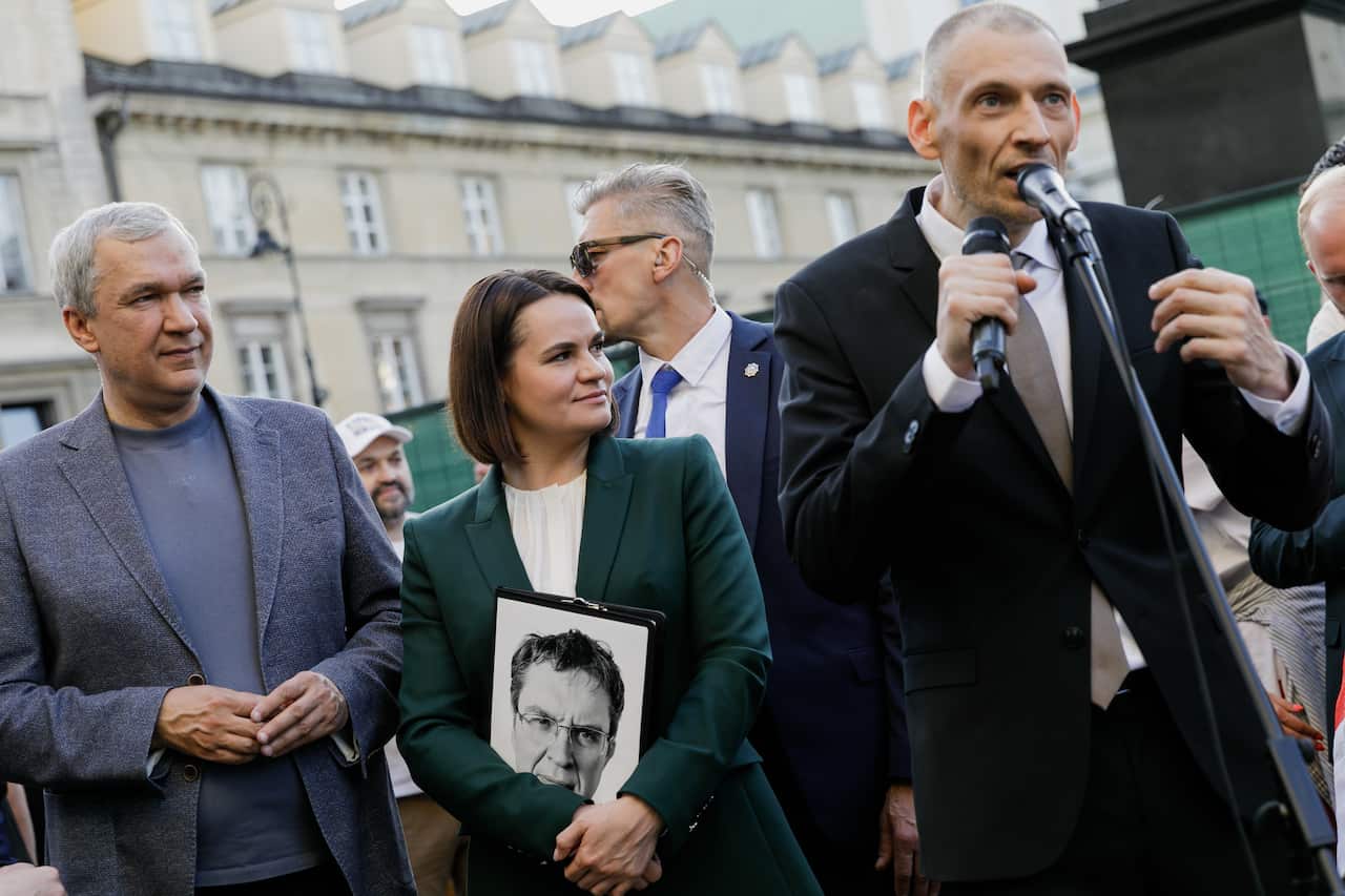 A woman wearing a dark green blazer and a white blouse stands to the right of a man wearing a black suit, speaking into a microphone.
