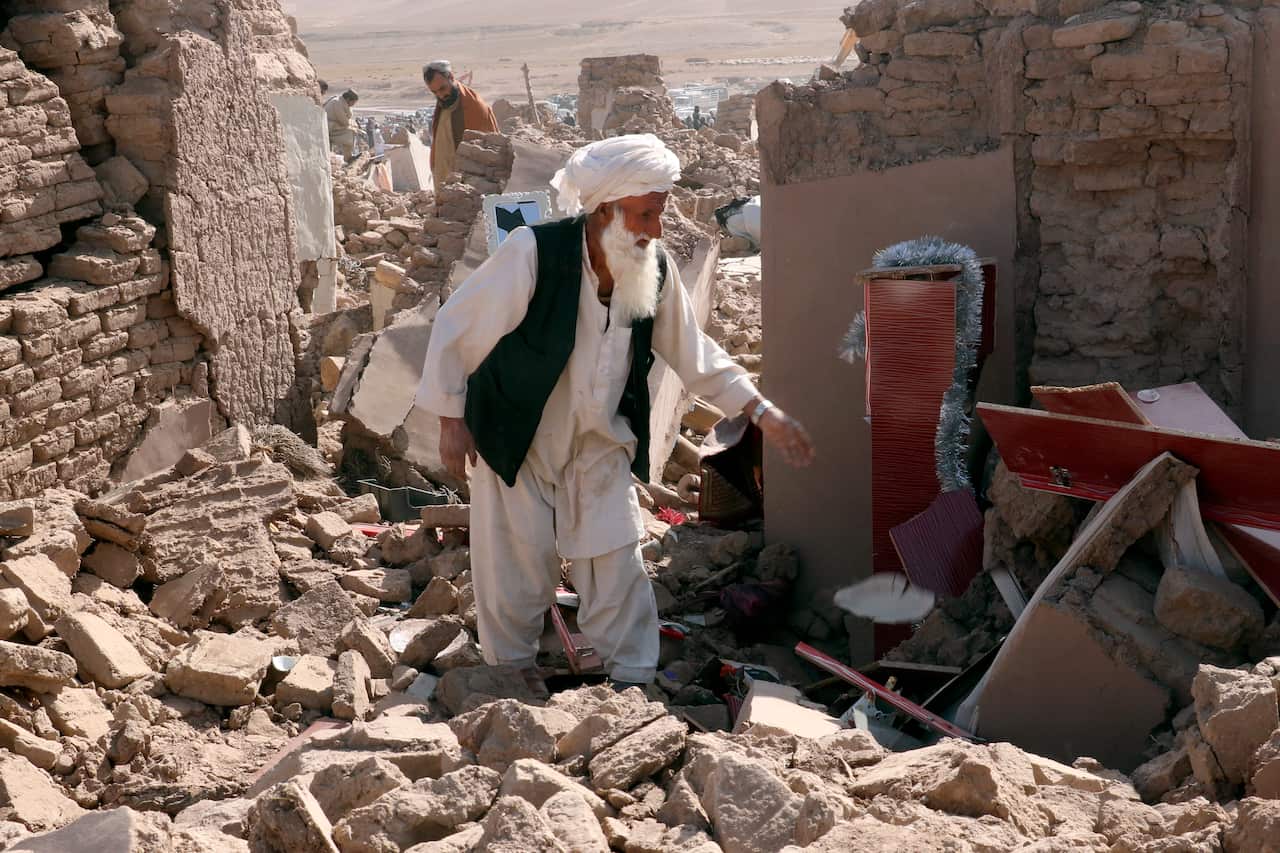 A bearded man wearing loose-fitting white pants and a long shirt walks through rubble. 