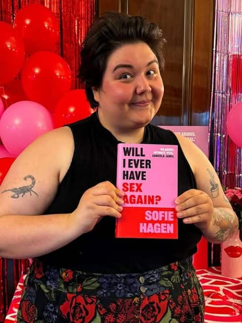A person in their 30s with short brown hair smiling while holding a red and pink book.