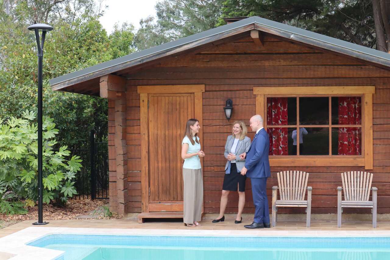 Three people stand talking between an outdoor pool and a wooden sauna building.