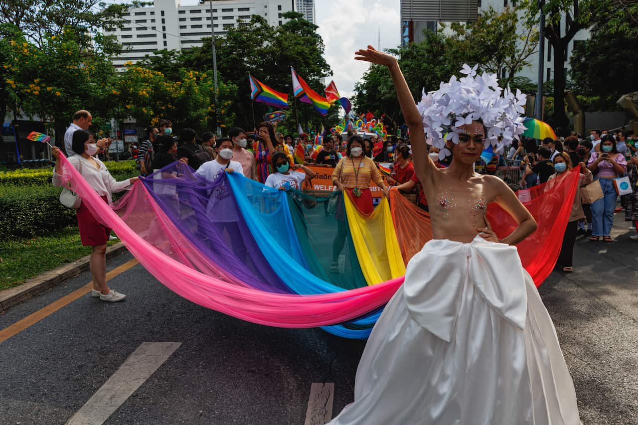 A person wearing a bridal gown with a rainbow-coloured train that several people are holding.