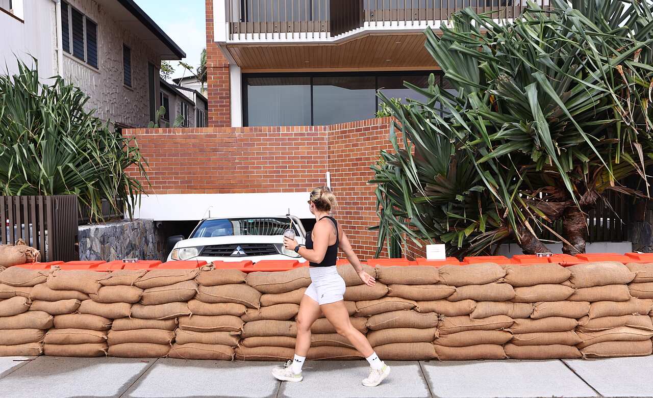 A woman in exercise clothes holding a drink walks past a row of sandbags outside a home