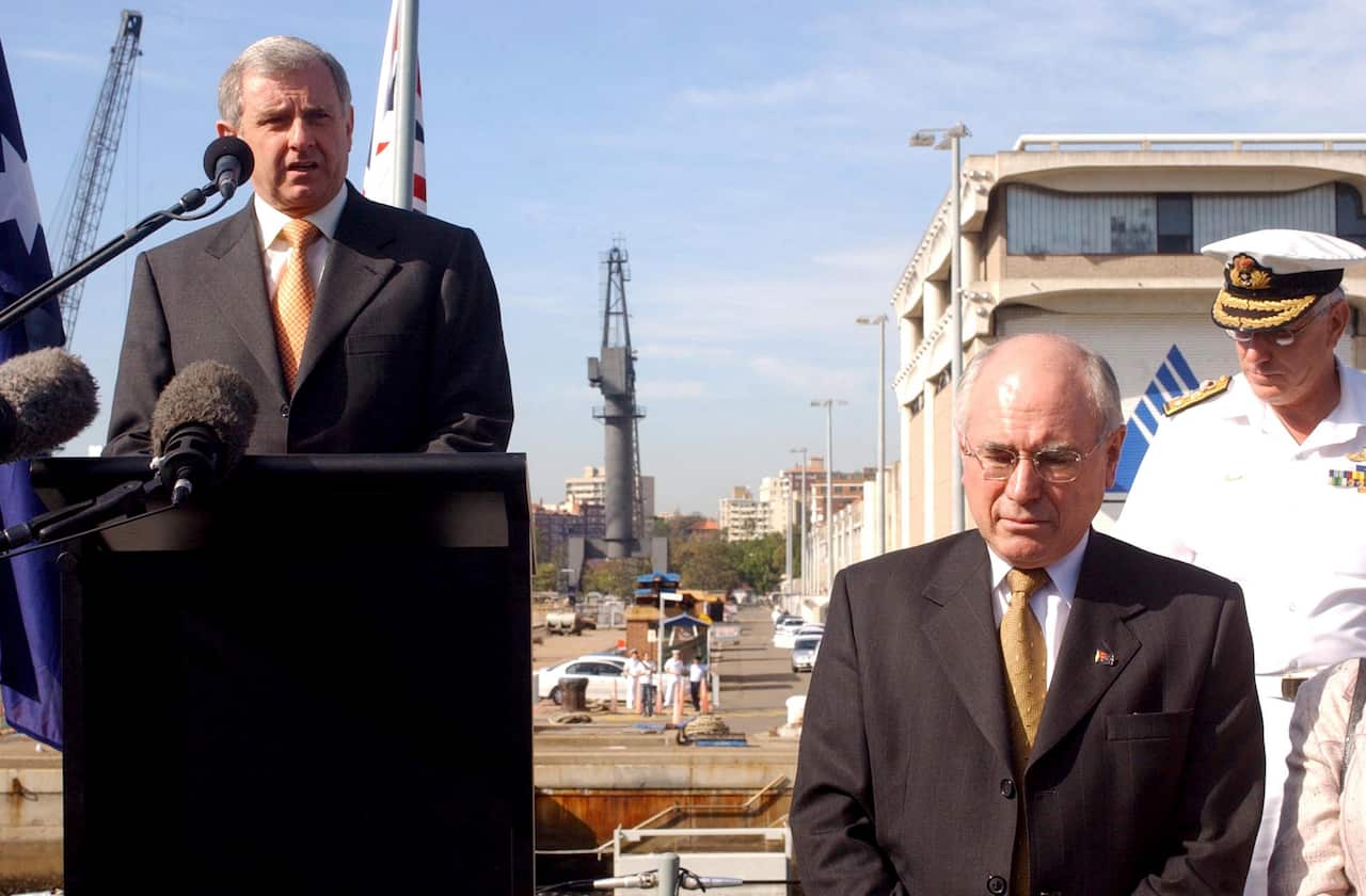A man in a suit talking from a raised lectern outside. There is another man in a suit standing below him to his left and another man in a white naval uniform