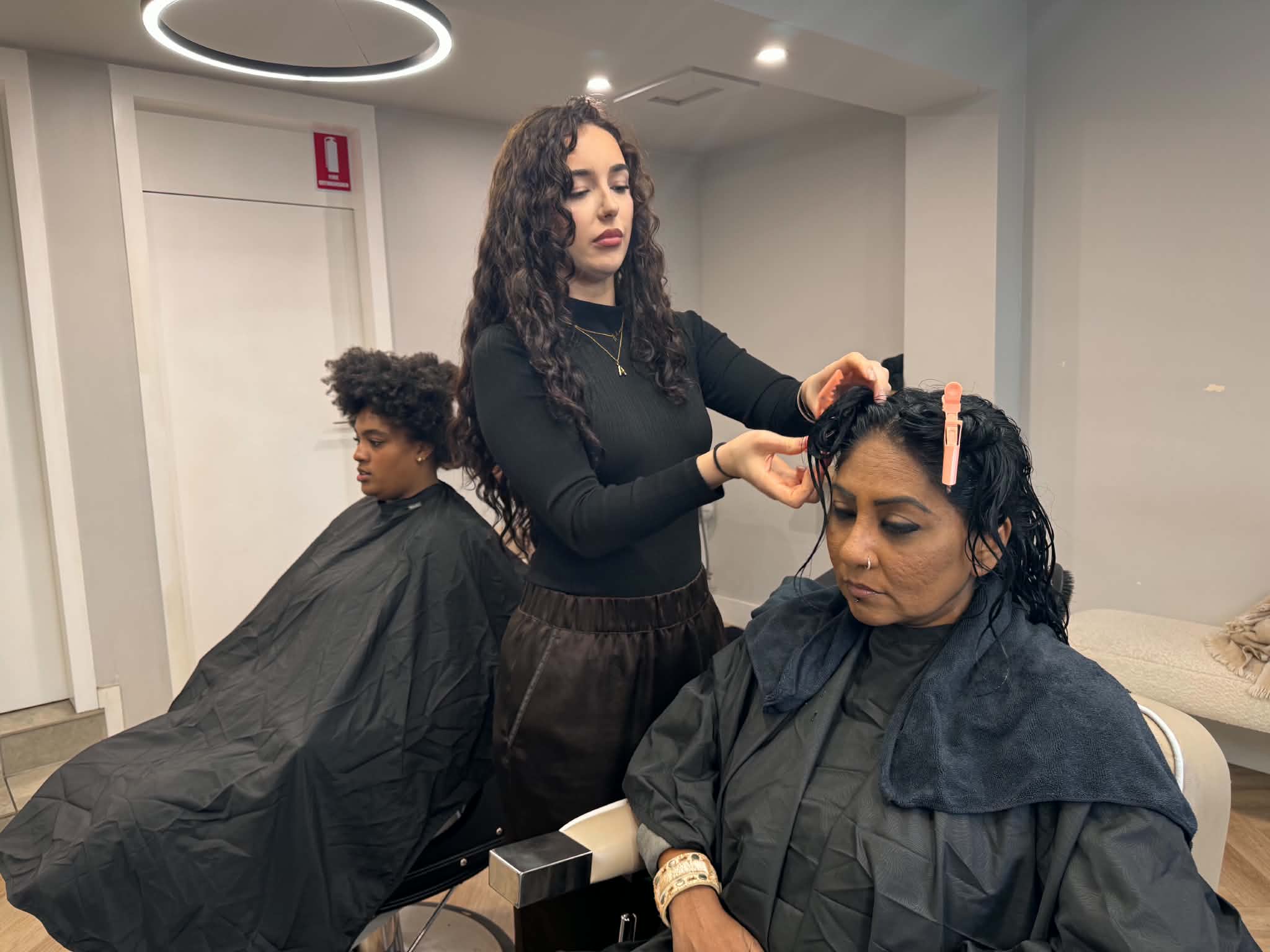 A hair stylist in an all-black outfit is working on a client's hair in a salon while another client with curly hair is sitting on a chair behind her.