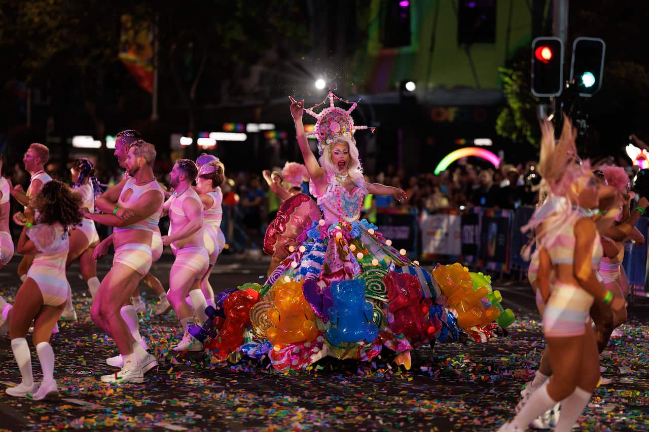A drag queen performs in the middle of a street covered in confetti. Her massive, colorful skirt is decorated with giant, translucent "gummy bears" and candy-themed ornaments. Dancers in pastel outfits surround her.