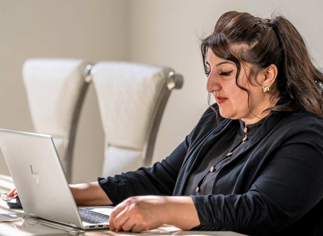 A woman with long brown hair in black clothing sits in front of a laptop.