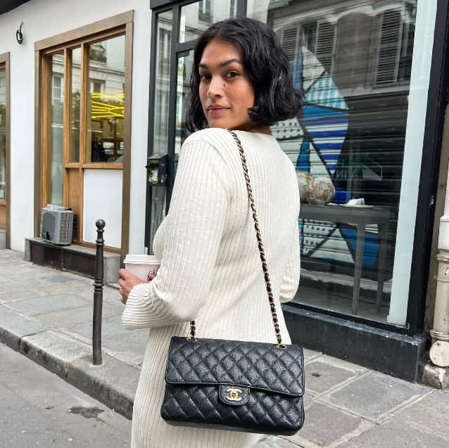 A woman with brown skin and black hair wearing a white dress holds a cup of coffee on a street. She's looking over her shoulder towards the camera.