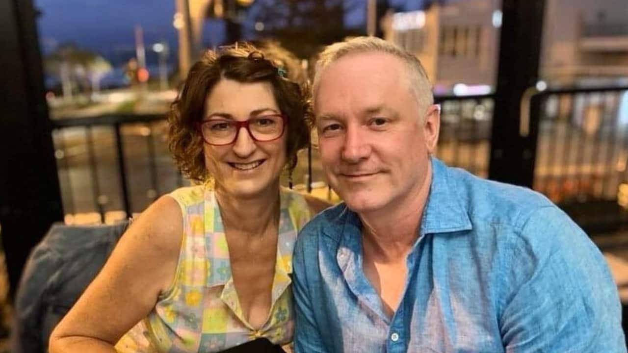 a middle aged woman and man smile at the camera, sitting close to each other on an outdoor balcony of a restaurant
