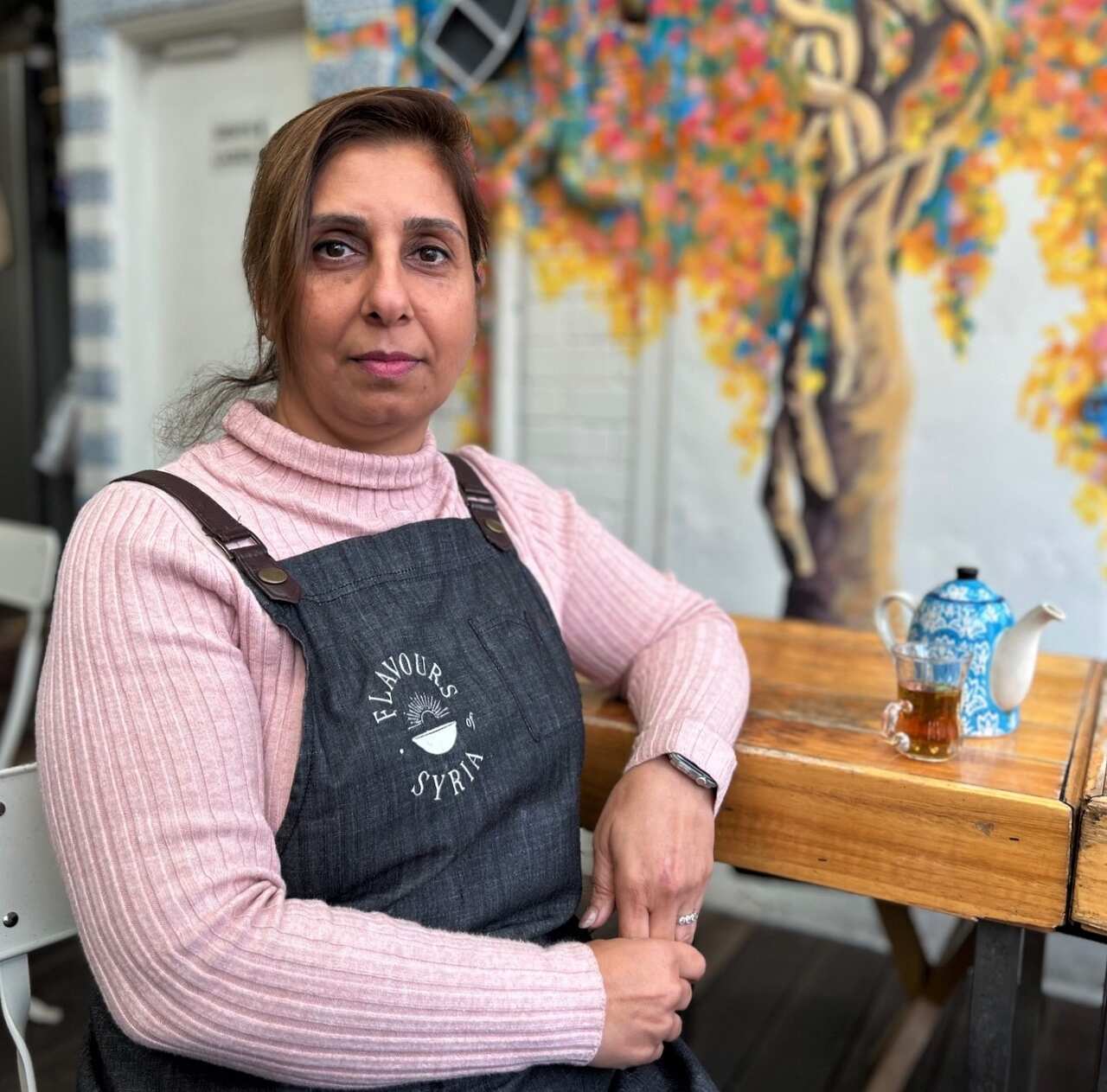 A woman in a pink jumper sits at a table with a serious expression.