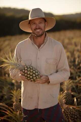 A man wearing a wide-brimmed hat in a field of pineapples, while holding a pineapple.