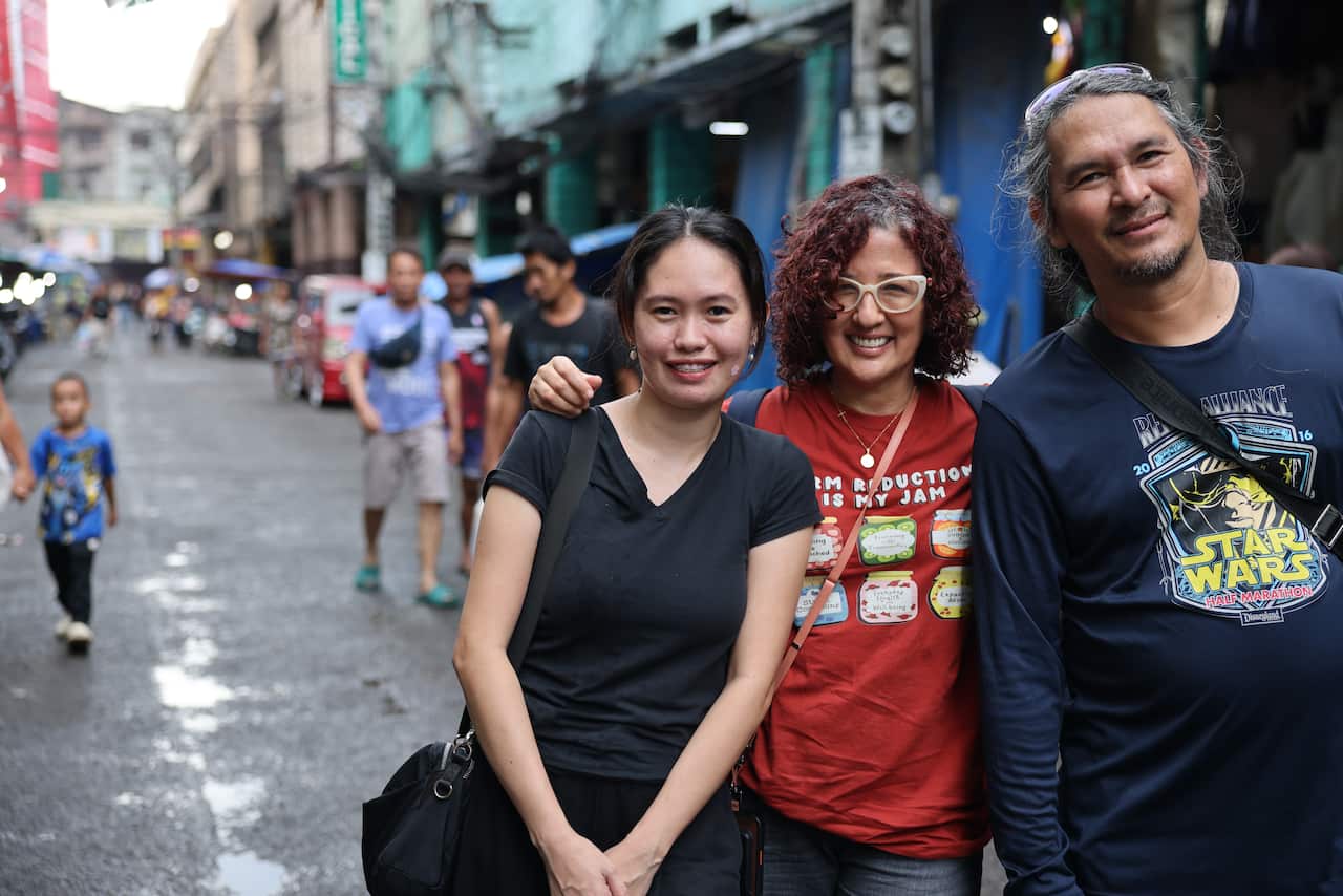 Three people stand in a street, smiling. There are people walking behind them. On the left is a young woman in a black t-shirt. The woman next to her has her arm around her and is wearing a red t-shirt with pictures of jam on it and text that reads 'harm reduction is my jam'. On the right is a smiling man in a long sleeve, dark blue Star Wars shirt. 