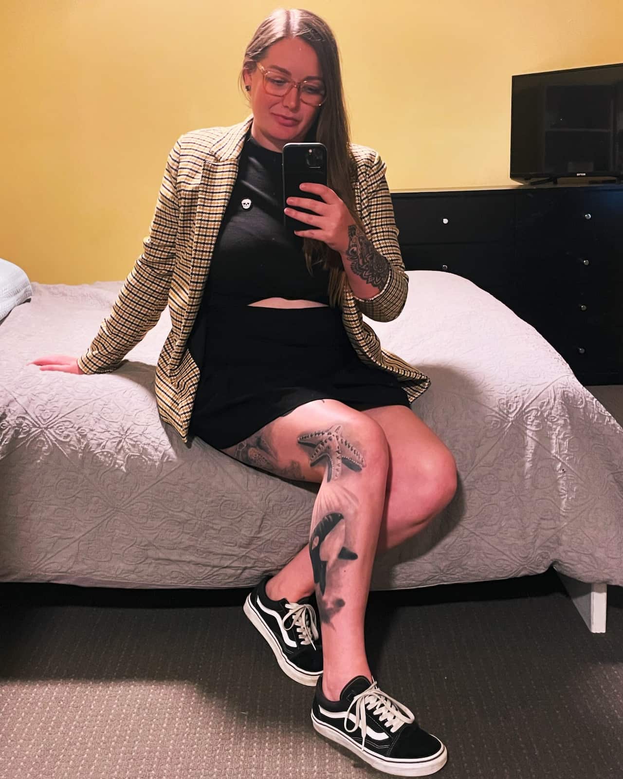 A woman in a tartan blazer and black dress with tattoos on her legs takes a mirror selfie on a bed