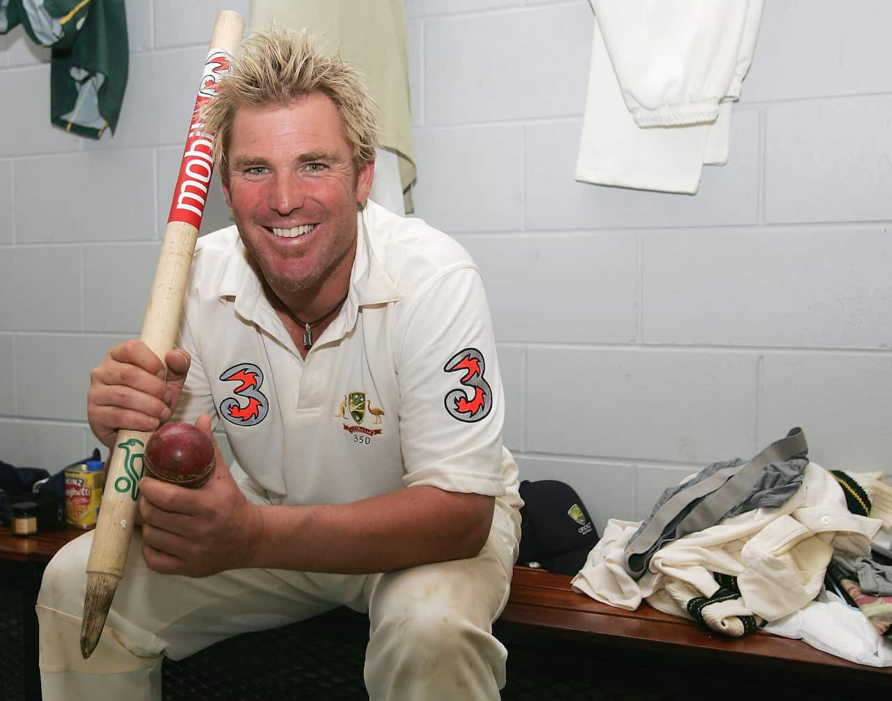 Shane Warne is seen sitting smiling as he sits on a bench while holding a stump and cricket ball.
