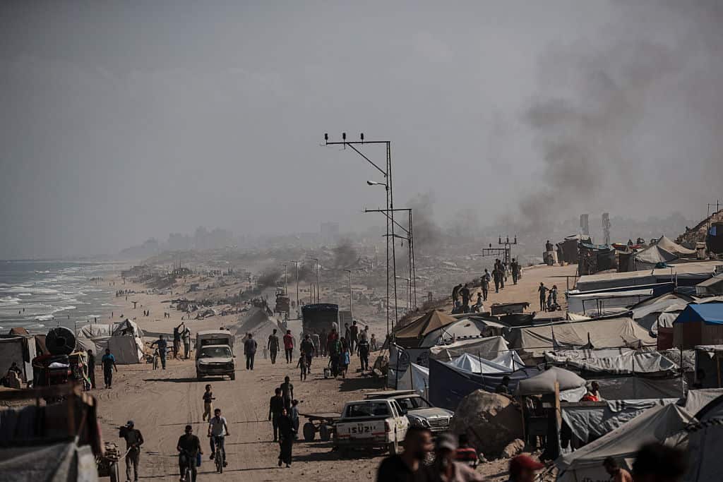 Tents housing displaced people line a street and a beach as smoke rises in the background.