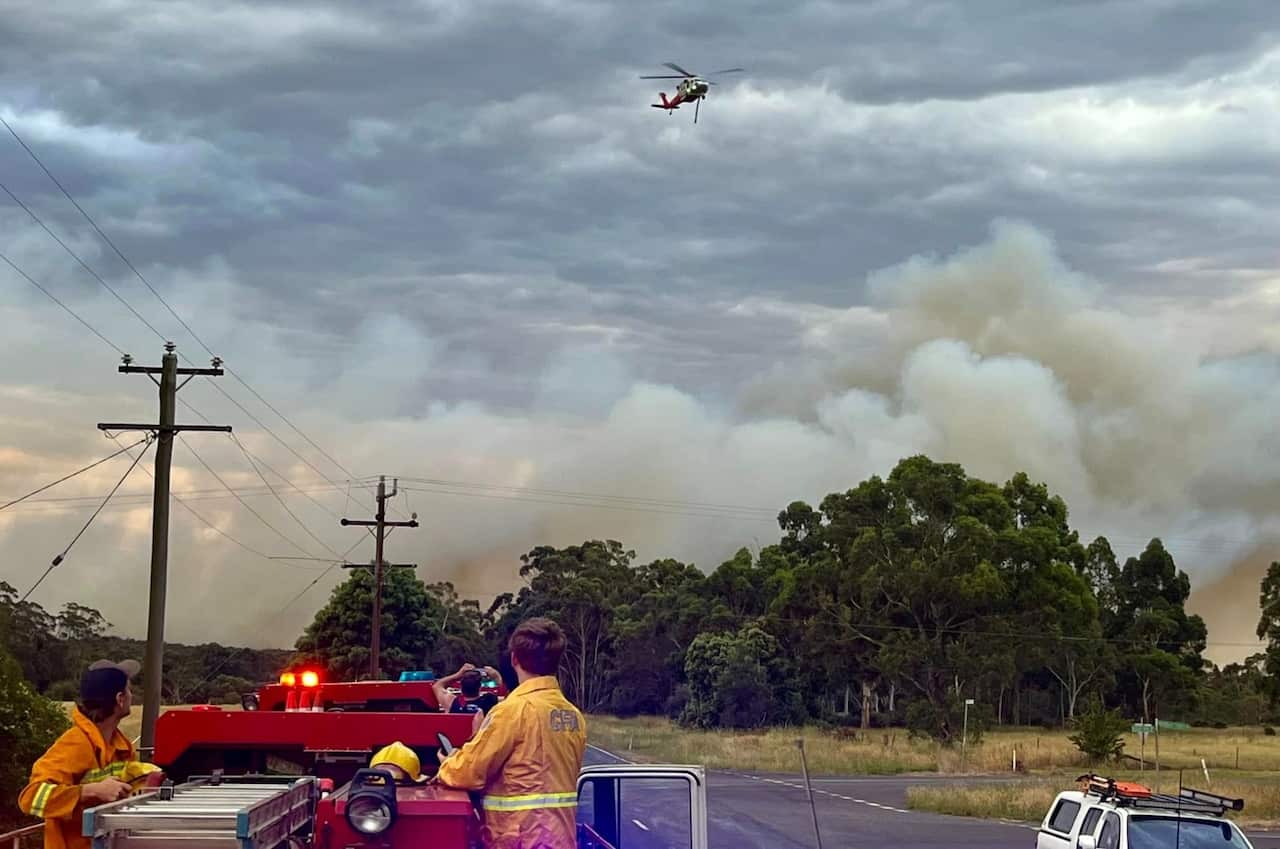 Two firefighters are standing on a fire truck, looking at the smoke billowing from a fire behind the trees. A helicopter can also be seen in the sky above.