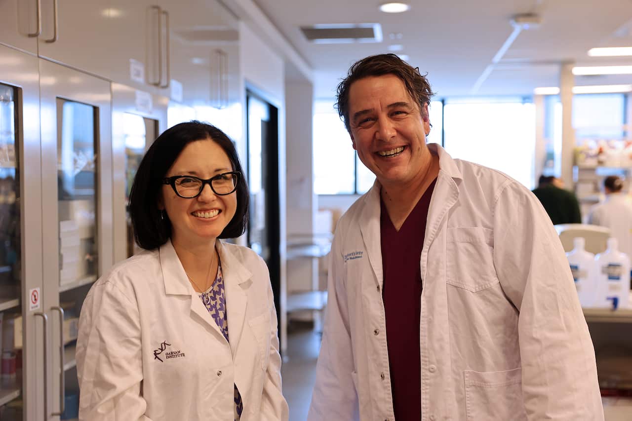 A man and a woman, both wearing white lab coats, stand in a laboratory. 