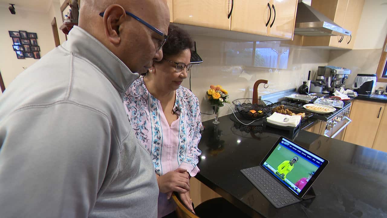 A man and a woman watching cricket on a laptop set on a kitchen bench