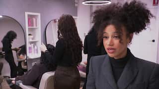A woman with curly hair is speaking in a salon with a hairstylist attending to a client behind her.