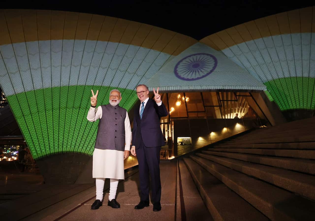 Narendra Modi and Anthony Albanese stand in front of the Sydney Opera House. The sails of the Opera House are illuminated with a light show using the colours of the Indian flag. Both leaders are making the peace gesture. 